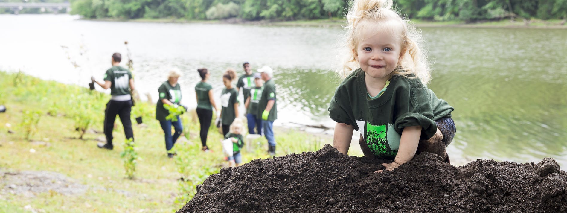 Little girl climbing on a pile of planting soil while volunteers plant trees